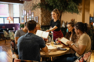 Waitress in a black top and red apron serves menus to four diners at a round wooden table in a cozy restaurant.